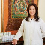 Asian lady standing next to a counter of Chinese herbal medicine with a Chinese green and orange mural in the background